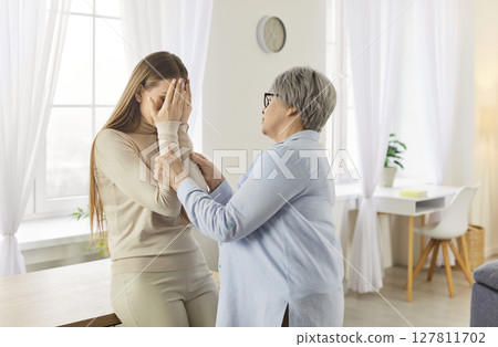 Senior mother supporting her worried unhappy adult child standing in living room at home together. Senior mother supporting her worried unhappy adult child standing in living room at home together. 127811702