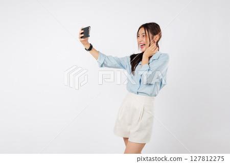 Happy young excited Asian woman laughing, taking a selfie picture with her smart mobile phone. She is making a quick selfie for social media. make video call isolated studio white background 127812275