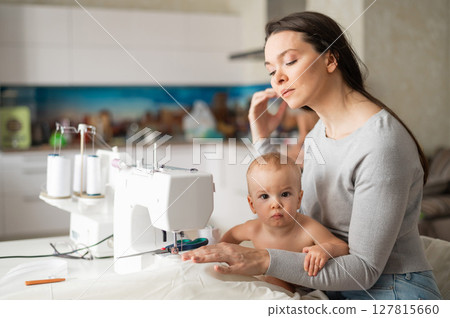 A young woman sews in the kitchen and holds a small child. Mom teaches her little son to sew on a sewing machine. Self-isolation homework A young woman sews in the kitchen and holds a small child. Mom teaches her little son to sew on a sewing machine. Self-isolation homework 127815660