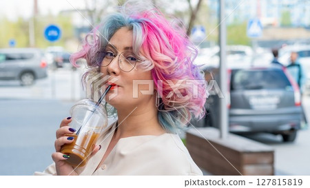 Close-up portrait of curly Caucasian woman with multi-colored hair wearing glasses. The hairstyle model is drinking a cold drink Close-up portrait of curly Caucasian woman with multi-colored hair wearing glasses. The hairstyle model is drinking a cold drink 127815819