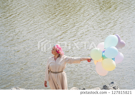 A woman with colored hair and an armful of balloons walks along the river embankment. 127815821
