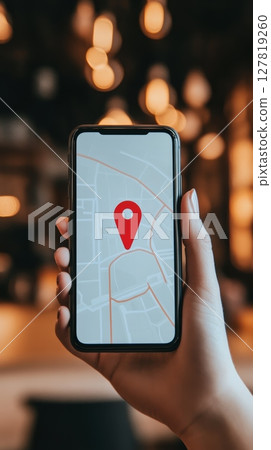 Hand holding a smartphone displaying a navigation app with a red location pin on a map, suggesting a search for directions or a point of interest in an out of focus background with warm lights 127819260
