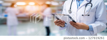 Doctor in white coat and stethoscope reviewing medical records in a busy hospital with blurred medical personnel, highlighting healthcare professionalism and patient care 127819261