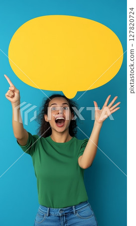 Excited young woman with curly hair points at a large empty yellow speech bubble while expressing enthusiasm and surprise against a vibrant blue background 127820724