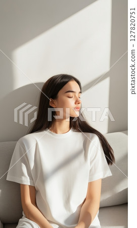Model wearing white t shirt sitting on sofa with closed eyes, enjoying sunlight streaming through window, creating peaceful and serene atmosphere, perfect for lifestyle and fashion campaign 127820751