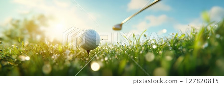 Golf club poised to strike a ball resting on the tee, surrounded by lush grass on a dewy golf course at sunrise, with sunlight filtering through trees in the background 127820815