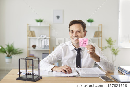 Happy man sitting on workplace in office holding a sticker with a light bulb drawn on it Happy man sitting on workplace in office holding a sticker with a light bulb drawn on it 127821718