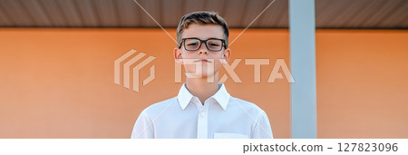 Portrait of a confident and intelligent young student wearing glasses and a crisp white shirt, standing against the backdrop of a modern school building, ready for the academic challenges ahead 127823096