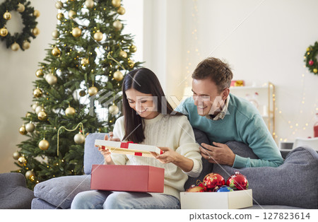 Happy young woman sitting on sofa at home with boyfriend and opening red present gift box. 127823614
