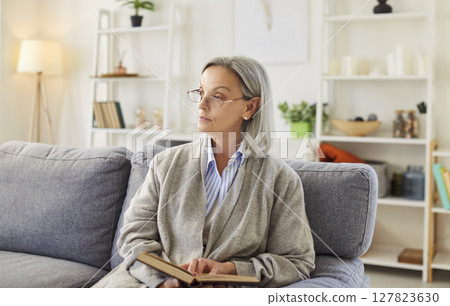 Portrait of senior elderly woman in eyeglasses sitting on sofa at home reading book in living room Portrait of senior elderly woman in eyeglasses sitting on sofa at home reading book in living room 127823630