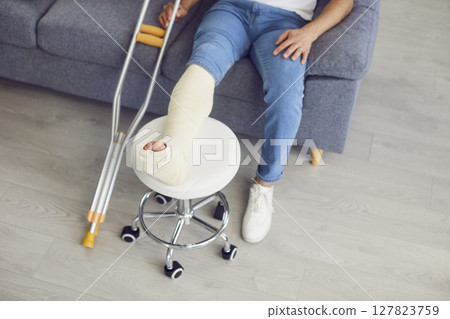 Man sitting on sofa at home on rehabilitation with crutches and leg in plaster cast after injury Man sitting on sofa at home on rehabilitation with crutches and leg in plaster cast after injury 127823759