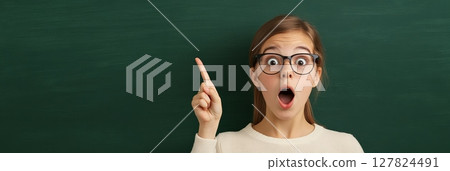 Smart student wearing eyeglasses pointing index finger up at empty blackboard with surprised face expression having idea during lesson in school Smart student wearing eyeglasses pointing index finger up at empty blackboard with surprised face expression having idea during lesson in school 127824491