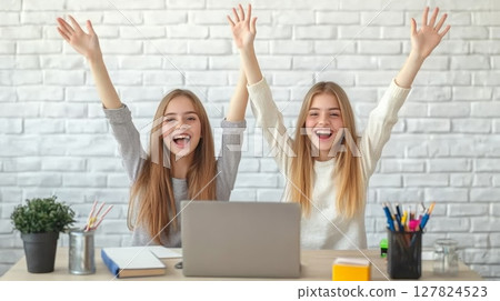 Two cheerful twin sisters students are sitting at desk with laptop, books and pencils, raising arms and celebrating successful online education or test results 127824523