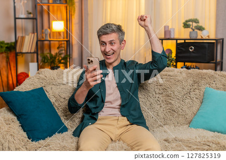 Smiling man celebrates smartphone victory with clenched fist, joyfully reacting to good result Smiling man celebrates smartphone victory with clenched fist, joyfully reacting to good result 127825319