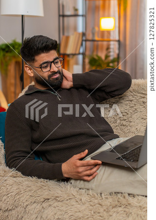 Indian sad man massaging neck on sofa with laptop at home, tired from long hours of screen work 127825321