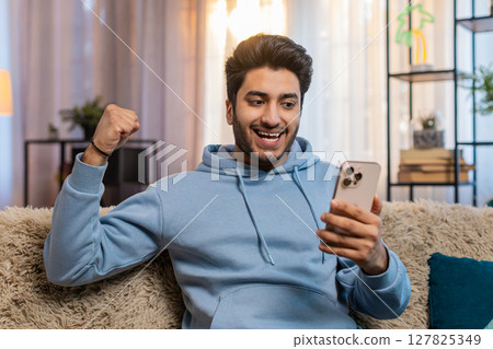Excited Indian man holding smartphone celebrating online game win sitting on sofa in living room Excited Indian man holding smartphone celebrating online game win sitting on sofa in living room 127825349