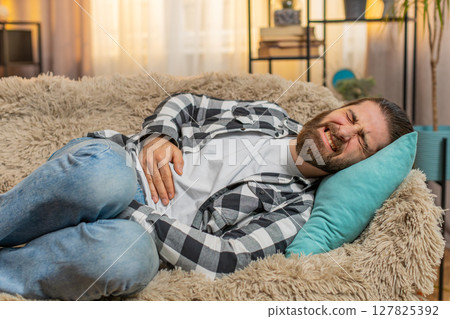 Young ill sick man lying at sofa with palm on abdomen moaning from stomach trouble or overeating Young ill sick man lying at sofa with palm on abdomen moaning from stomach trouble or overeating 127825392