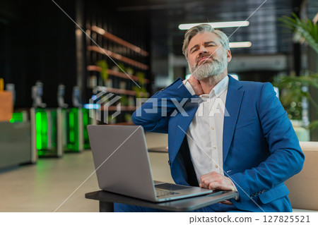 Tired middle-aged businessman suffers from spine neck muscle pain working on laptop in office lobby 127825521