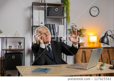 Overjoyed senior businessman sitting at home office desk listening to music through headphones Overjoyed senior businessman sitting at home office desk listening to music through headphones 127825581