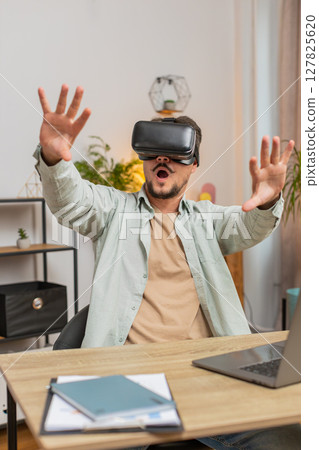 Confident young Caucasian man freelancer using VR headset while sitting at home office table desk Confident young Caucasian man freelancer using VR headset while sitting at home office table desk 127825620
