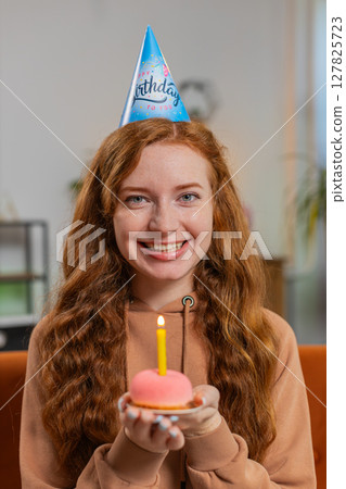Happy woman wears birthday hat makes wish congratulating blowing burning candle on cake at home Happy woman wears birthday hat makes wish congratulating blowing burning candle on cake at home 127825723