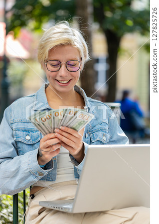 Happy rich young woman using laptop winner victory holding cash money in dollar banknotes in city 127825726