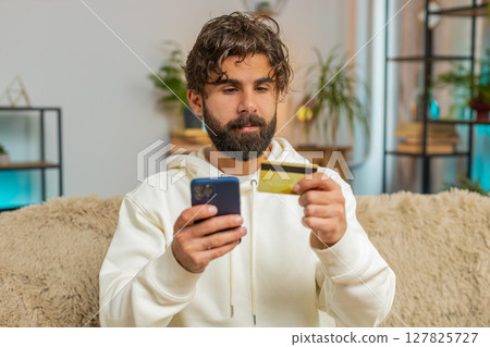 Happy young Hispanic man guy using credit bank card and cellphone while shopping online at home Happy young Hispanic man guy using credit bank card and cellphone while shopping online at home 127825727