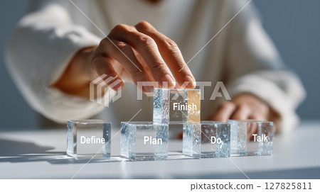 Person arranging translucent glass blocks with words like Define, Plan, Do, Talk, and Finish, symbolizing project management 127825811