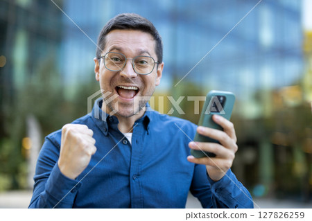 A man joyfully reacts to something he sees on his phone, fist clenched in celebration while outside. 127826259