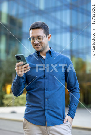 A man in glasses is using a smartphone outside in front of a modern office building, he seems pleased. A man in glasses is using a smartphone outside in front of a modern office building, he seems pleased. 127826261