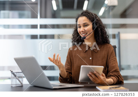 A smiling woman wearing a headset engages in a video call in an office setting, holding a tablet. A smiling woman wearing a headset engages in a video call in an office setting, holding a tablet. 127826353