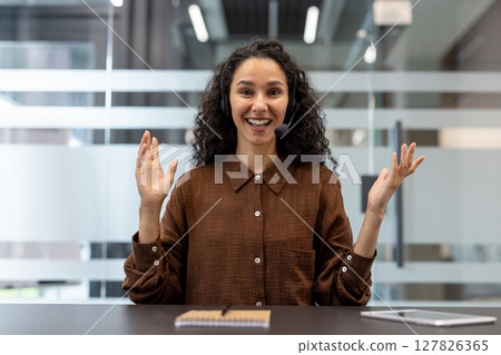 A smiling woman wearing a headset gestures while sitting at a desk in a modern office, communicating. 127826365