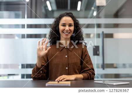 A smiling woman with curly hair waves at the camera while wearing a headset in an office setting. 127826367