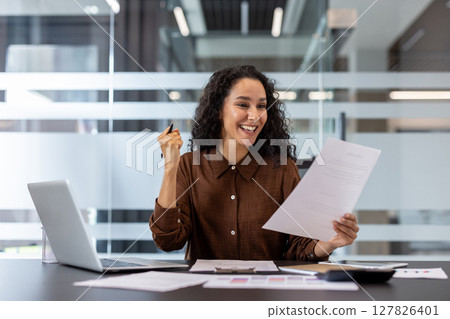 A joyful woman in an office celebrates a success, holding a document and smiling brightly. She is sitting in front of a laptop. 127826401