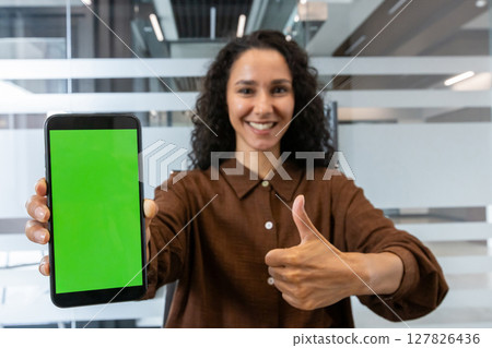 A woman is holding a smartphone with a green screen, smiling and giving a thumbs-up gesture, possibly in an office setting. 127826436