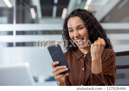 A joyous woman celebrates good news on her phone, fist raised in triumph with a beaming smile. A joyous woman celebrates good news on her phone, fist raised in triumph with a beaming smile. 127826466