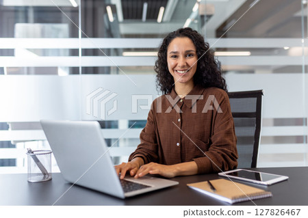 A smiling young woman works at a laptop in a modern office, radiating confidence and professionalism. She is surrounded by office supplies. 127826467