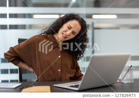 A woman with curly hair experiences back pain while working on her laptop at an office desk. She is visibly in discomfort, holding her back. 127826473