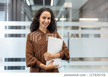 A smiling woman holds a laptop in an office setting, possibly a businesswoman or employee. A smiling woman holds a laptop in an office setting, possibly a businesswoman or employee. 127826726