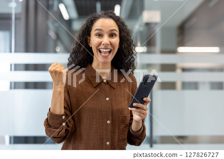 An excited woman celebrates a win while looking at her phone. She appears elated and is holding her phone in one hand. An excited woman celebrates a win while looking at her phone. She appears elated and is holding her phone in one hand. 127826727
