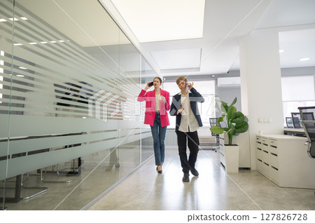 Two colleagues are walking through the office and talking on the phones. Two colleagues are walking through the office and talking on the phones. 127826728