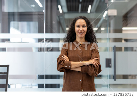 A smiling woman stands confidently in an office setting, arms crossed, radiating professionalism and approachability. 127826732