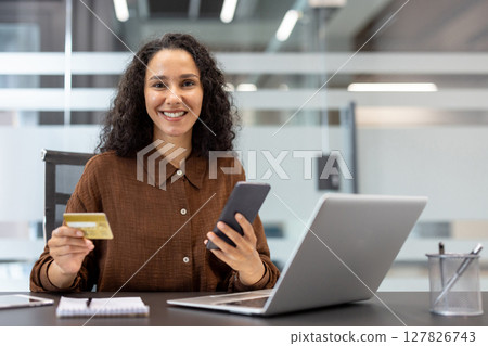A smiling woman in a brown shirt holds a credit card and smartphone while seated at her desk with a laptop. A smiling woman in a brown shirt holds a credit card and smartphone while seated at her desk with a laptop. 127826743