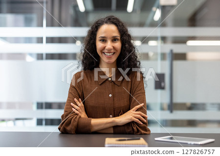 A smiling businesswoman in a modern office setting. She has curly hair and is wearing a brown shirt, her arms crossed. A smiling businesswoman in a modern office setting. She has curly hair and is wearing a brown shirt, her arms crossed. 127826757
