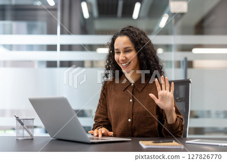 Young successful businesswoman using laptop for video call. Company employee inside office smiling cheerfully greeting, waving hand, online business conference. Young successful businesswoman using laptop for video call. Company employee inside office smiling cheerfully greeting, waving hand, online business conference. 127826770