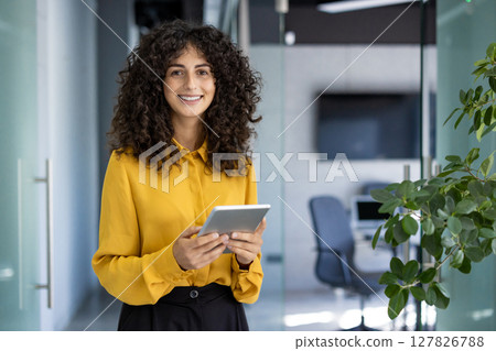 Hispanic businesswoman stands confidently in modern office holding tablet. She wears yellow shirt, smiling brightly, conveying professionalism, success, and positivity in work environment. Hispanic businesswoman stands confidently in modern office holding tablet. She wears yellow shirt, smiling brightly, conveying professionalism, success, and positivity in work environment. 127826788