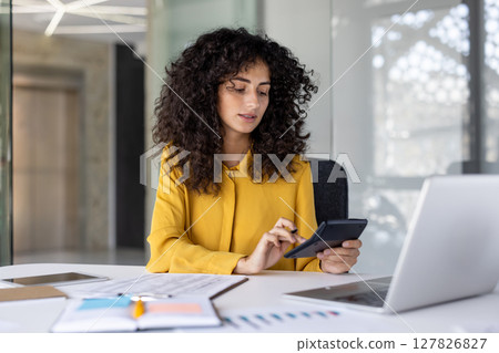 A focused woman works diligently at a desk, utilizing a calculator alongside a laptop and financial documents in a bright office. 127826827