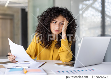 A woman with curly hair, wearing a yellow shirt, reviews documents, looking tired while working on her laptop in the office setting. A woman with curly hair, wearing a yellow shirt, reviews documents, looking tired while working on her laptop in the office setting. 127826875