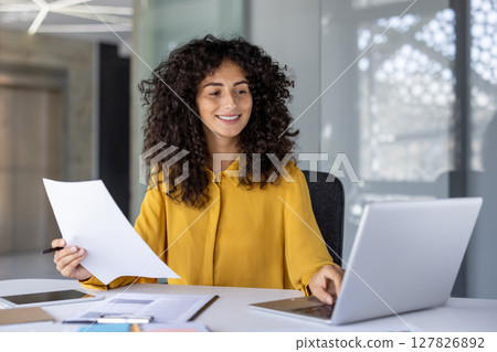 Hispanic businesswoman smiling reviewing documents and working on laptop in office. Engaged and focused, she represents confidence and professionalism in workplace, showing successful career woman Hispanic businesswoman smiling reviewing documents and working on laptop in office. Engaged and focused, she represents confidence and professionalism in workplace, showing successful career woman 127826892