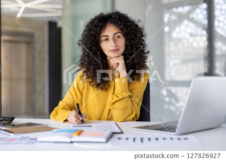 Serious and confident businesswoman working with documents and reports. Woman thinking about current financial investment tasks, preparing report, and filling tax forms while sitting inside office. 127826927